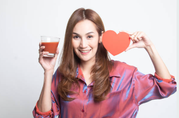 Smiling person holding a glass of reddish-brown juice and a red heart cutout, symbolizing love, wellness, and heart health