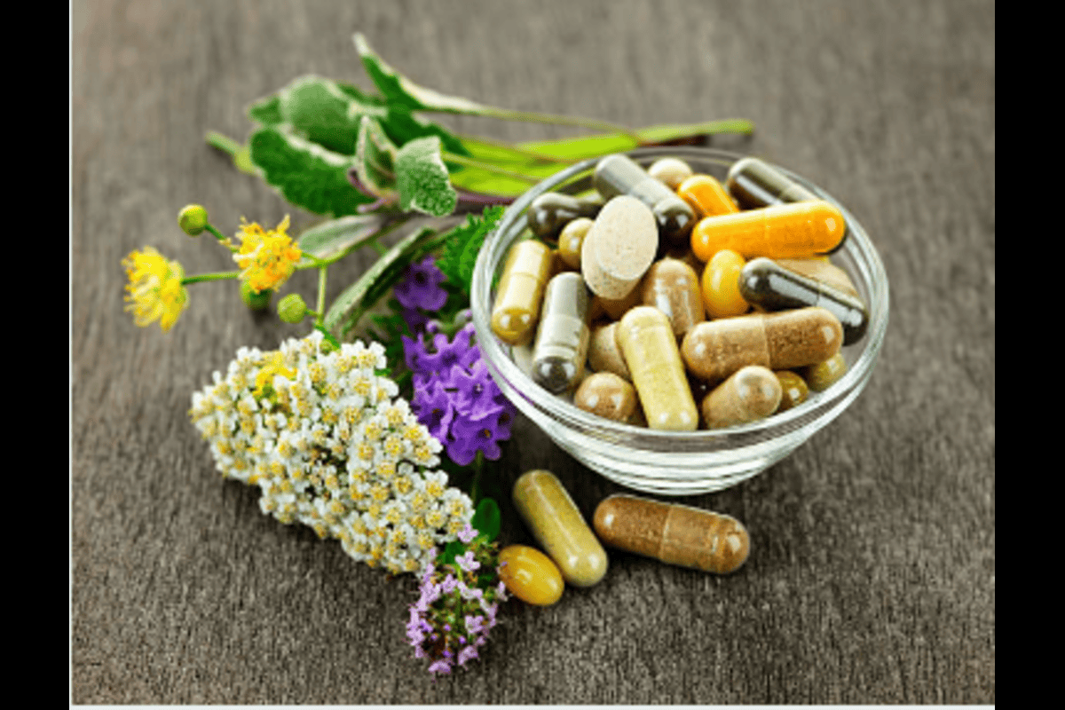 White bowl of assorted dietary supplements surrounded by fresh herbs and colorful flowers on wooden surface, representing holistic health and natural medicine