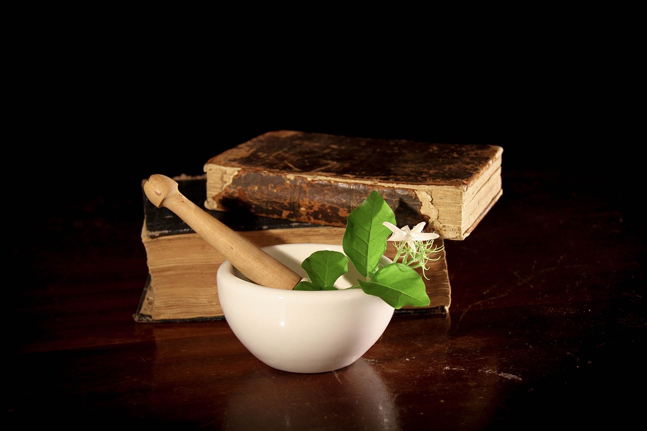 Image of a white ceramic mortar and pestle containing fresh green leaves and a small white flower, placed on a dark wooden surface with an aged herbal book in the background—symbolizing traditional medicine, natural healing, and ancient Ayurvedic wisdom