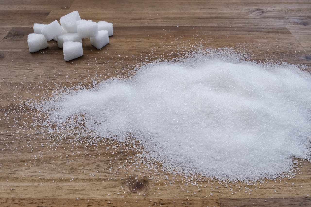 Close-up image of granulated white sugar spread across a wooden surface, with a small stack of white sugar cubes in the background—highlighting the contrast between loose and compact forms of sugar used in culinary and nutritional contexts